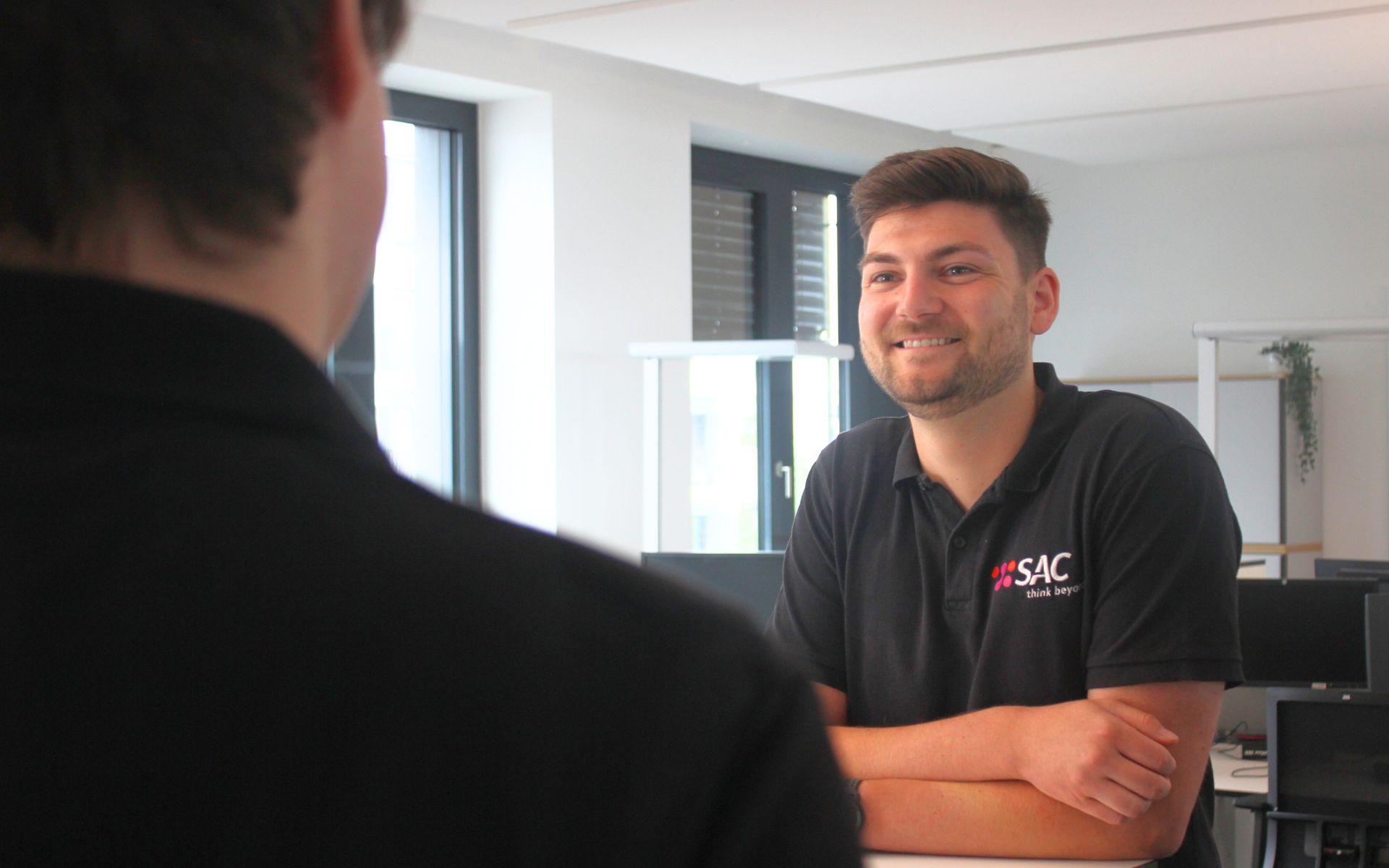 Lächelnder Mitarbeiter des IT-Service Provider SAC im Büro in einem schwarzen SAC Polohemd spricht zu einer anderen Person
