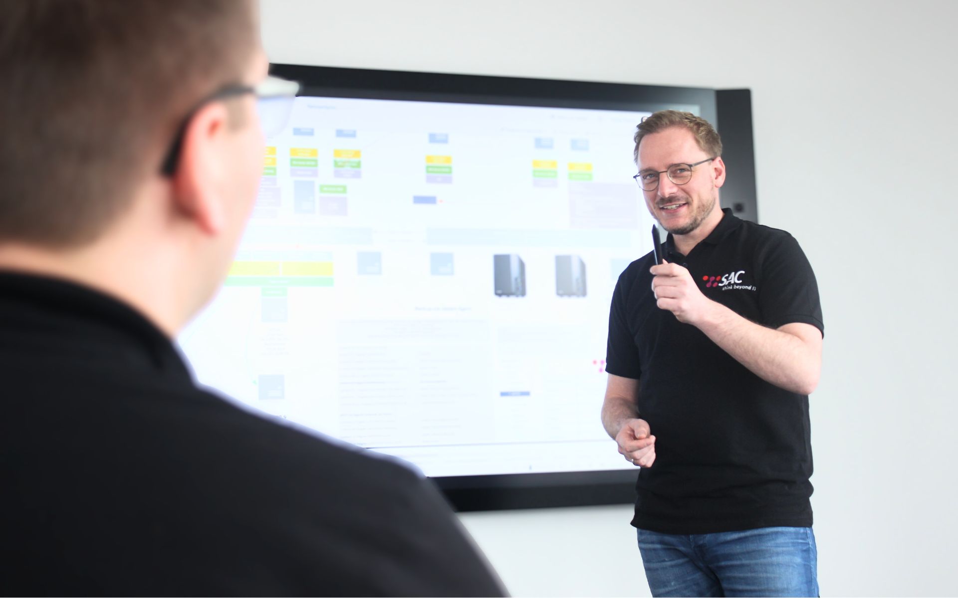 Auf dem Foto sieht man zwei SAC Mitarbeiter in einem Meetingraum. Der eine Mitarbeiter erläutert den anderen gerade eine Bestandsdokumentation der IT-Infrastruktur eines Kunden.